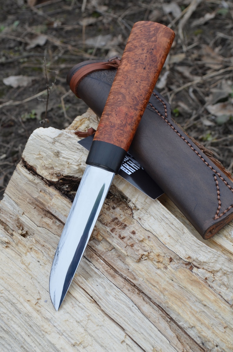 Puede incluir: Un cuchillo artesanal con una hoja de acero pulido y un mango de madera de ra&iacute;z. El cuchillo descansa sobre un trozo de madera, con una funda de cuero marr&oacute;n en el fondo. El mango tiene una virola negra.