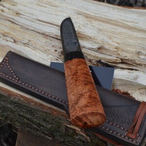 Puede incluir: Un cuchillo artesanal con una hoja oscura y un mango de madera de ra&iacute;z. El cuchillo descansa sobre una superficie de madera junto a una funda de cuero marr&oacute;n. El mango tiene un patr&oacute;n de vetas arremolinadas &uacute;nico.