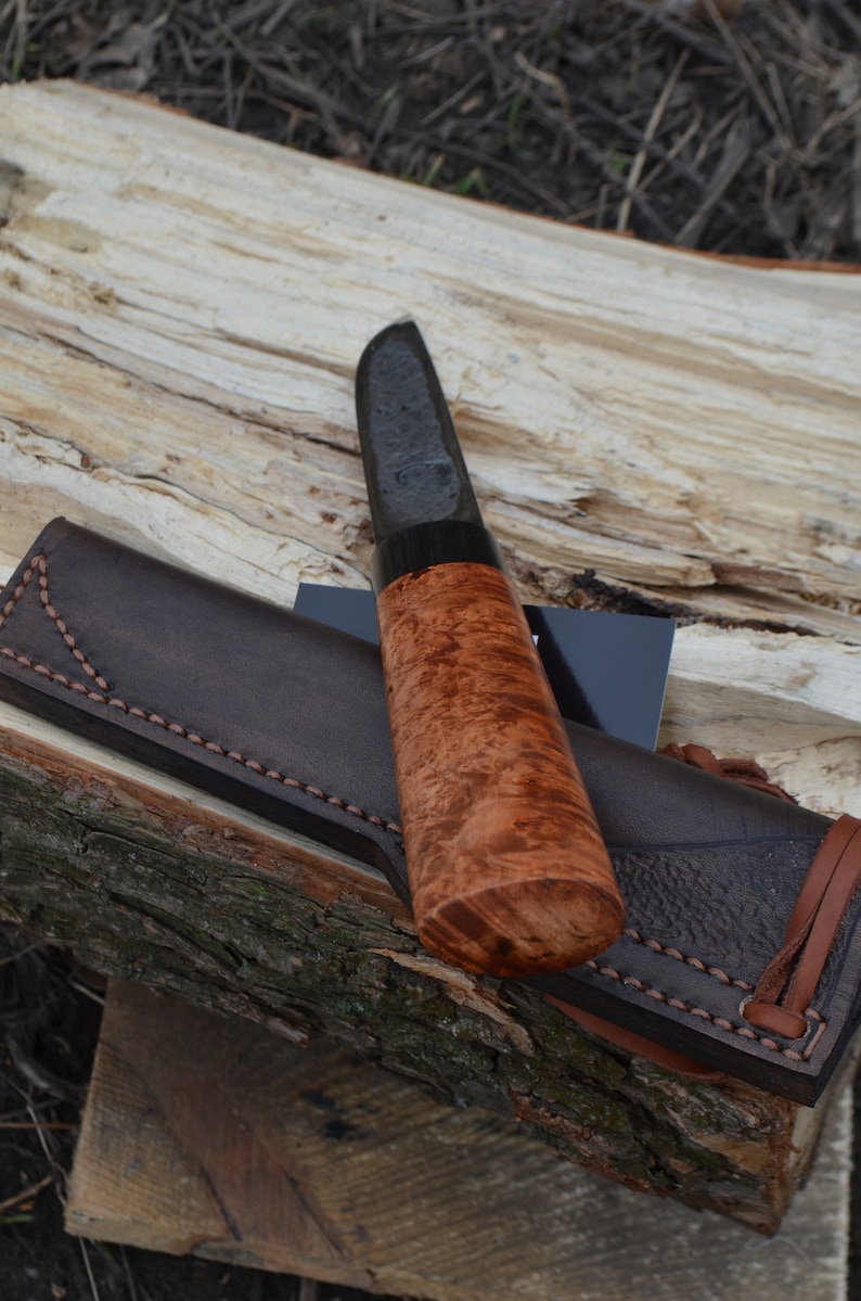 Puede incluir: Un cuchillo artesanal con una hoja oscura y un mango de madera de ra&iacute;z. El cuchillo descansa sobre una superficie de madera junto a una funda de cuero marr&oacute;n. El mango tiene un patr&oacute;n de vetas arremolinadas &uacute;nico.