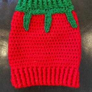 May include: A handmade crochet strawberry cozy. The cozy is primarily red with a green top and green drips. The bottom of the cozy has a ribbed design. The cozy is designed to fit over a bottle or container.