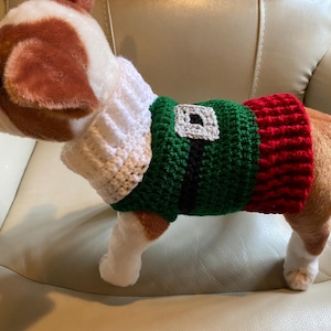 May include: A small dog wearing a crocheted elf costume. The costume is green, red, and white with a black belt. The dog is sitting on a white couch.