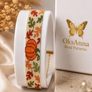 May include: A beaded bracelet featuring a cream background with a repeating pattern of orange pumpkins and multi-colored autumn leaves in shades of red, orange, and green. The bracelet has a gold-toned metal clasp and a thin gold chain adjustment.