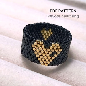 Può includere: Un anello nero di perline con un motivo a cuore dorato, realizzato con la tecnica del punto peyote. Il testo "PDF PATTERN Peyote heart ring" è visibile. Gioiello fatto a mano.