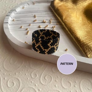 Könnte beinhalten: Ein schwarzer und goldener Perlenring mit geometrischem Muster. Der Ring liegt auf einer weißen Oberfläche mit dem Wort "PATTERN" auf einem lila Kreis.
