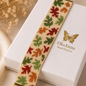 May include: A woven bead bracelet featuring a repeating pattern of autumn leaves in shades of red, orange, and green on a cream background. The bracelet has gold-toned metal end caps and a gold-toned chain extender. The bracelet rests on a white box.