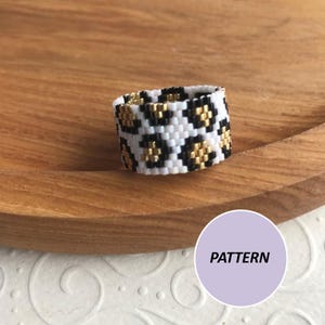 Può includere: Un anello di perline bianche, nere e dorate con un motivo a stampa leopardata. L'anello è su una superficie di legno. PATTERN.