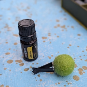 May include: A small brown essential oil bottle with a yellow label that reads "Lemon" and a green felt ball car air freshener with a black clip. The items are on a blue surface with gold paint splatters.