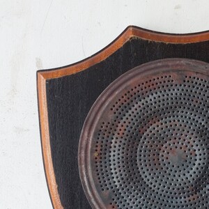 May include: An antique metal heat diffuser with a black handle, mounted on a shield-shaped wooden plaque. The plaque has a black painted surface with a brown wooden border. The diffuser has a circular design with numerous small holes.