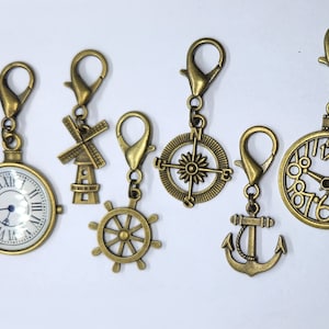 May include: Six antique gold-toned charms with different designs, including a compass, a windmill, a clock, a ship's wheel, and an anchor. Each charm has a lobster claw clasp.