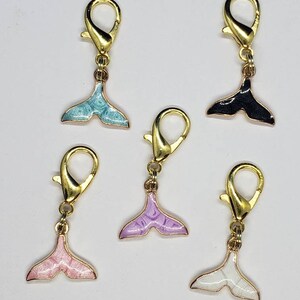 May include: Five gold-tone metal whale tail charms with different colored enamel finishes: light blue, black, lavender, pink, and white.