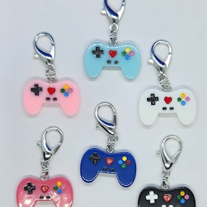 May include: Six colorful plastic video game controller charms with silver keychains. The controllers are in blue, pink, white, black, and two shades of pink. Each controller has a red heart, a black plus sign, and colorful circles.