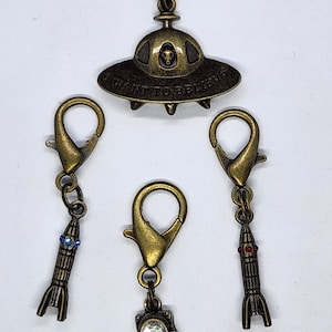 May include: Set of four gold-tone metal charms featuring an astronaut, two rockets, and a UFO with the text "I Want To Believe".