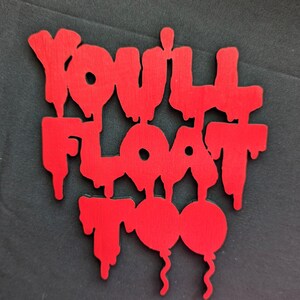 May include: A red wooden sign with the phrase "YOU'LL FLOAT TOO" in a dripping font. Two red balloons are at the bottom of the sign. The sign is on a black background.