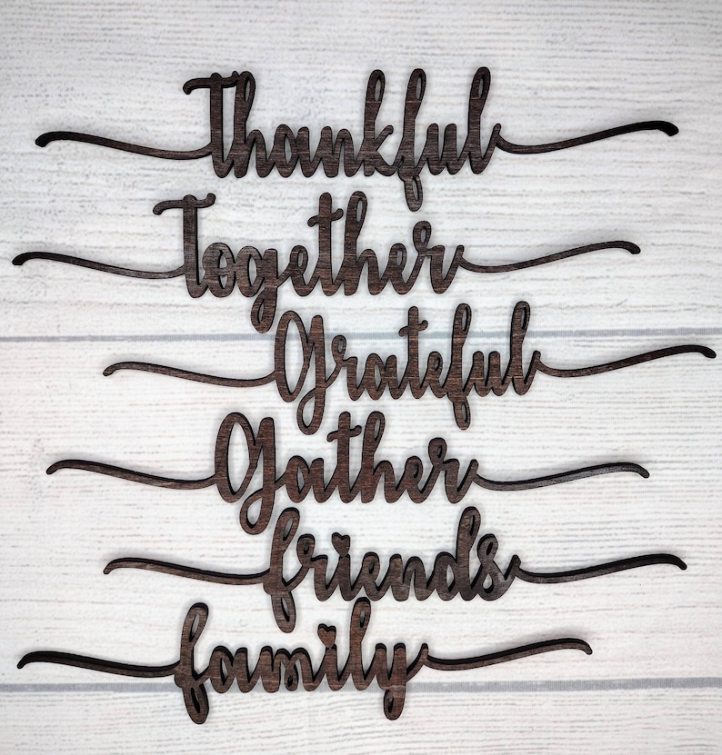 May include: Wooden cutout words spelling "Thankful", "Together", "Grateful", "Gather", "Friends", and "Family" on a white wood background.