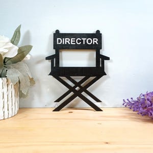 May include: A black wooden director's chair with the word "DIRECTOR" in white lettering. The chair is set against a light blue background and rests on a wooden surface. Decorative flowers are in the background.