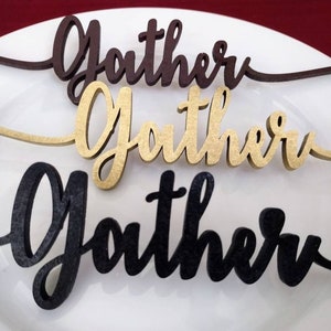 May include: Three wooden "Gather" signs in different colors: brown, gold, and black. The signs are on a white plate.