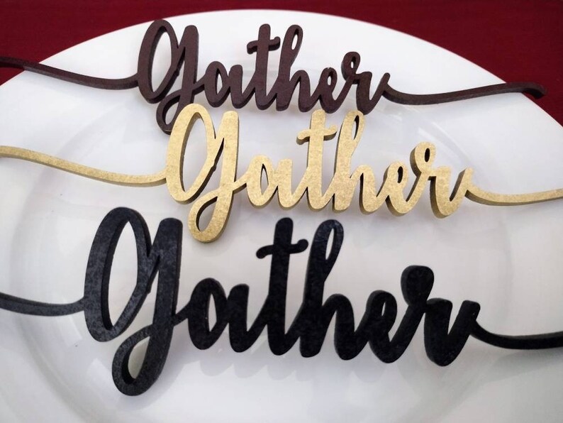 May include: Three wooden "Gather" signs in different colors: brown, gold, and black. The signs are on a white plate.