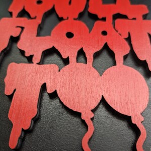 May include: Red wooden sign with the words "You'll Float Too" in a stylized font. The sign features a balloon design. The sign is set against a black background.