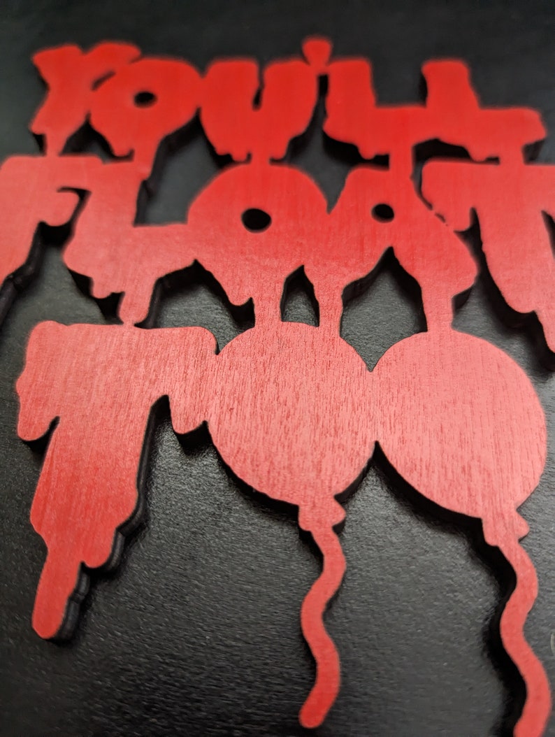 May include: Red wooden sign with the words "You'll Float Too" in a stylized font. The sign features a balloon design. The sign is set against a black background.