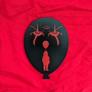 Puede incluir: Globo negro con una silueta roja de una cara de payaso y un niño sosteniendo un globo rojo. Los ojos del payaso son grandes y la boca es un ceño fruncido.