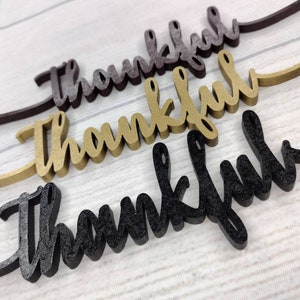 May include: Three wooden signs with the word "thankful" written in cursive. The signs are in different colors: black, gold, and brown.