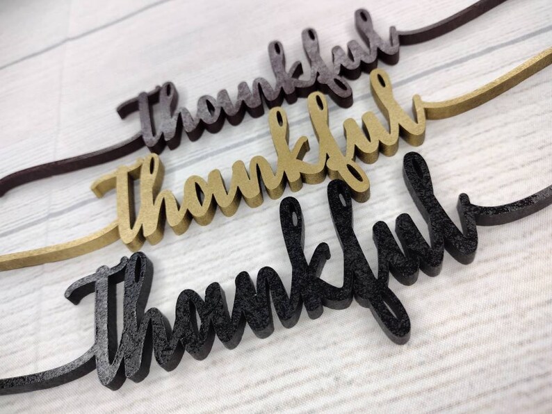 May include: Three wooden signs with the word "thankful" written in cursive. The signs are in different colors: black, gold, and brown.