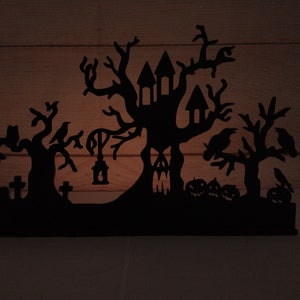 May include: Black silhouette of a haunted house with a spooky tree, crows, and tombstones. The house has a pointed roof and windows. The tree has bare branches and is surrounded by crows. The tombstones are simple and have crosses on top. The silhouette is cut out of wood and is a Halloween decoration.