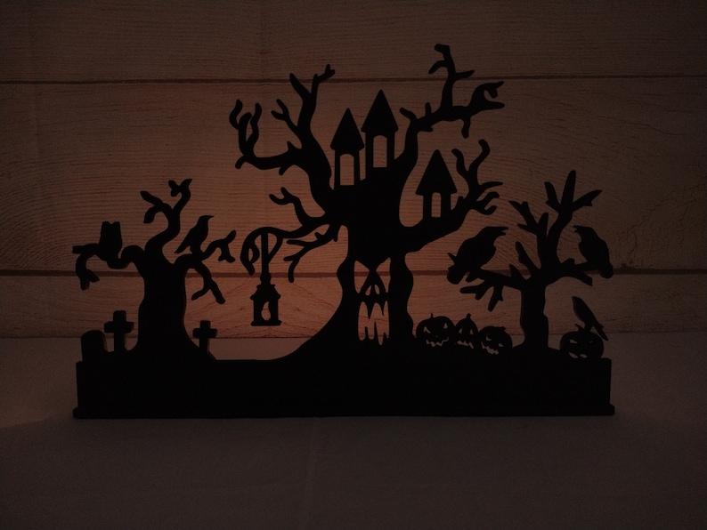 May include: Black silhouette of a haunted house with a spooky tree, crows, and tombstones. The house has a pointed roof and windows. The tree has bare branches and is surrounded by crows. The tombstones are simple and have crosses on top. The silhouette is cut out of wood and is a Halloween decoration.