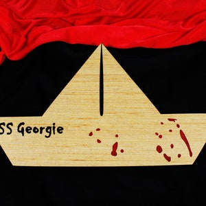 Puede incluir: Un recorte de madera con forma de barco con el texto "SS Georgie" y salpicaduras rojas, sobre un fondo negro con tela roja drapeada. El barco es de madera clara.