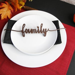 May include: A white dinner plate with a black napkin on top. The plate has a wooden sign that says "family" on it. The plate is on a red placemat and a black tablecloth. There is a small orange pumpkin on the table.