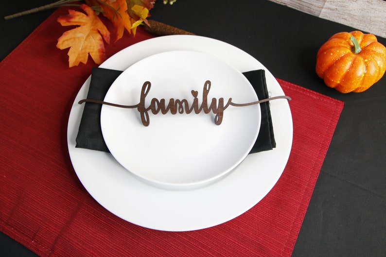 May include: A white dinner plate with a black napkin on top. The plate has a wooden sign that says "family" on it. The plate is on a red placemat and a black tablecloth. There is a small orange pumpkin on the table.