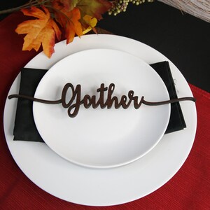 May include: A white plate with a black napkin on top. A wooden place card with the word "Gather" is placed on top of the napkin. The plate is on a red placemat.