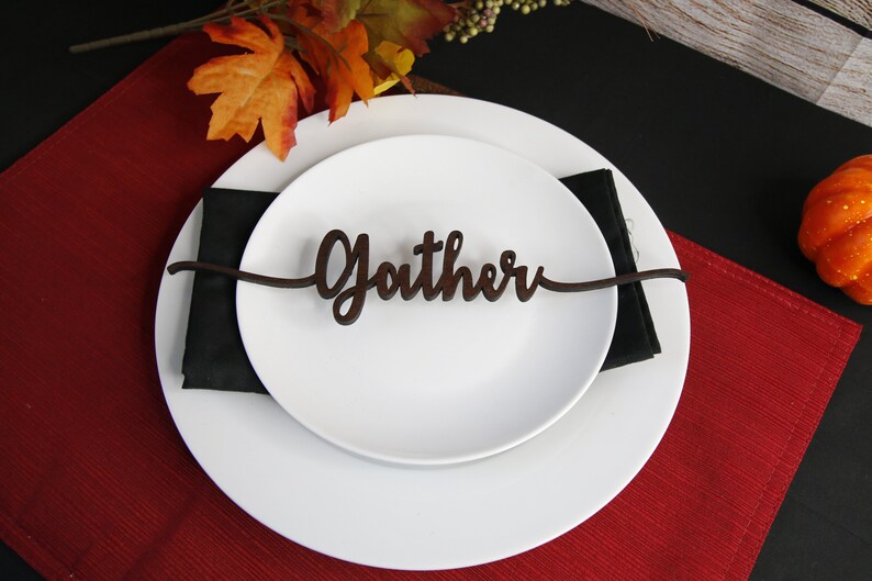 May include: A white plate with a black napkin on top. A wooden place card with the word "Gather" is placed on top of the napkin. The plate is on a red placemat.