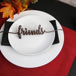 May include: A white dinner plate with a black napkin and a brown wooden "Friends" place card on a red placemat. A small orange pumpkin sits on a black surface.
