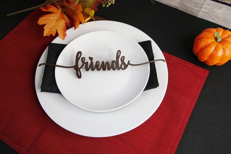 May include: A white dinner plate with a black napkin and a brown wooden "Friends" place card on a red placemat. A small orange pumpkin sits on a black surface.