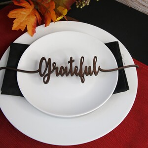 May include: A white plate with a brown wooden place card that says "Grateful" on it. The plate is on a red placemat and a black napkin.