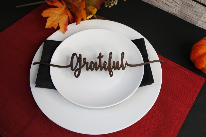 May include: A white plate with a brown wooden place card that says "Grateful" on it. The plate is on a red placemat and a black napkin.