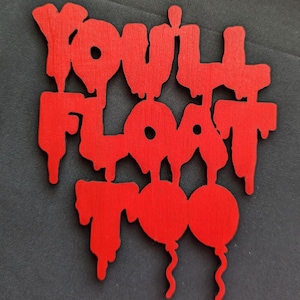 May include: A red wooden cutout with the phrase "You'll Float Too" in a dripping font. Two red balloon shapes are at the bottom. The cutout is on a dark gray surface.