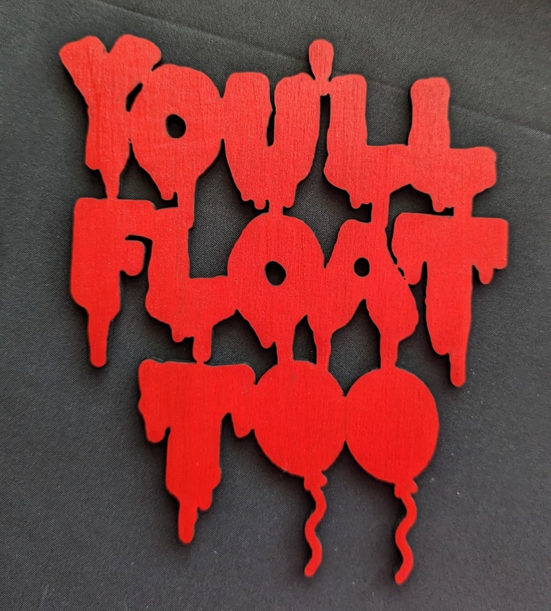 May include: A red wooden cutout with the phrase "You'll Float Too" in a dripping font. Two red balloon shapes are at the bottom. The cutout is on a dark gray surface.