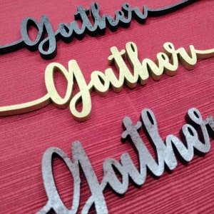 May include: Three wooden signs with the word "Gather" written in different styles. The top sign is black, the middle sign is gold, and the bottom sign is brown.