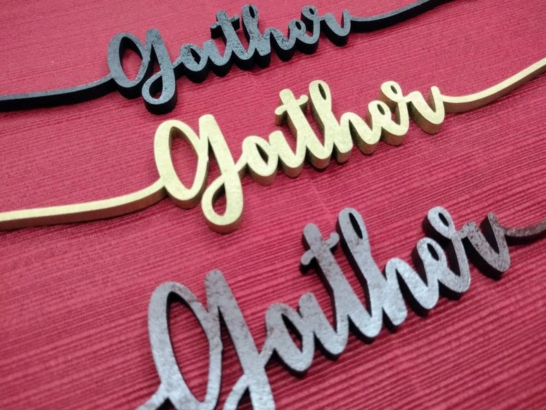 May include: Three wooden signs with the word "Gather" written in different styles. The top sign is black, the middle sign is gold, and the bottom sign is brown.