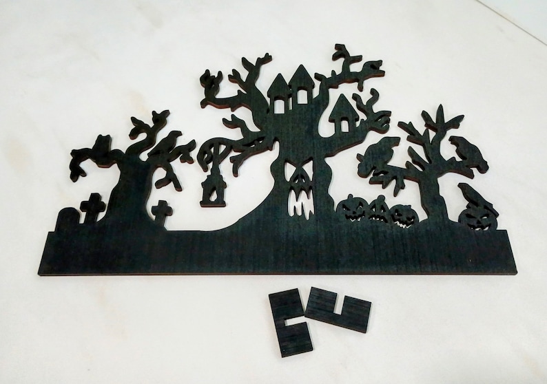 May include: Black wooden Halloween silhouette of a haunted house with trees, pumpkins, and a graveyard. The silhouette is cut out of a single piece of wood.