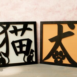 Op de afbeelding: Twee zwarte houten borden met uitgesneden Chinese karakters. Het bord aan de linkerkant heeft de karakters "猫" en "田" en een zwart katten silhouet. Het bord aan de rechterkant heeft het karakter "大" en een potafdruk silhouet. Beide borden hebben een lichte achtergrond.