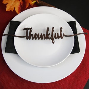 May include: A white plate with a black napkin on top. A wooden place card with the word "Thankful" is placed on top of the napkin. The plate is on a red placemat.