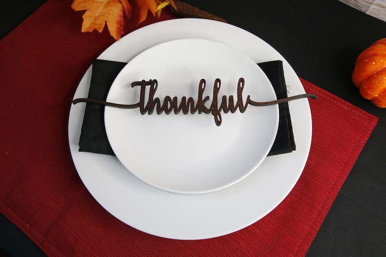 May include: A white plate with a black napkin on top. A wooden place card with the word "Thankful" is placed on top of the napkin. The plate is on a red placemat.