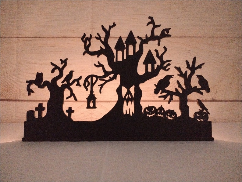 May include: Black silhouette of a haunted house, trees, and pumpkins. The house is a treehouse with a spooky face carved into the trunk. There are crows perched on the branches. This is a wooden Halloween decoration.