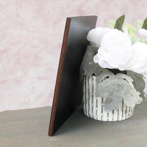 Op de afbeelding: Een donkerbruin houten bord dat tegen een witte plantenbak met witte bloemen en groene bladeren leunt.