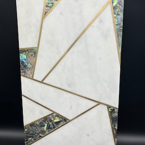 Op de afbeelding: Een rechthoekig decoratief stuk van wit marmer met een geometrisch ontwerp. Het ontwerp bevat goudkleurige lijnen en iriserende abalone schelp accenten. Het stuk heeft een gepolijste afwerking en een moderne esthetiek.