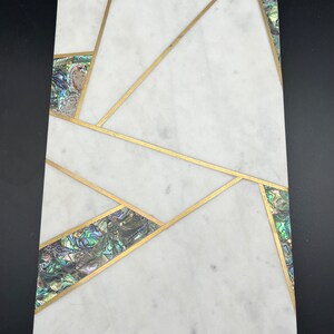 Op de afbeelding: Rechthoekig wit marmeren serveerplank met een geometrisch ontwerp met gouden inlegwerk. Secties van iriserende abalone schelp zijn gezet in de gouden lijnen. De plank is ongeveer 46 cm lang en 20 cm breed.