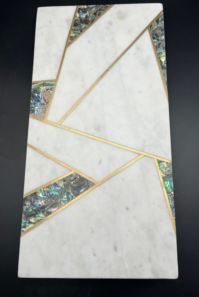 Op de afbeelding: Rechthoekig wit marmeren serveerplank met een geometrisch ontwerp met gouden inlegwerk. Secties van iriserende abalone schelp zijn gezet in de gouden lijnen. De plank is ongeveer 46 cm lang en 20 cm breed.